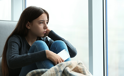 depression-children-v1 Upset teenage girl with smartphone sitting at window indoors.